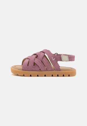 Rožinės zomšos šlepetės su kryžminiu dirželiu, paminkštintu vidpadžiu ir guminėmis padais, kuriuose yra zigzago raštas, užtikrinantis sukibimą. Reguliuojamas kulno dirželis.