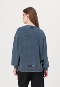 Modra denim majica s kapuco, ki se ponaša z ohlapnim krojem, rebrastim ovratnikom in manšetami, vidnimi šivi ter poudarki na stranskem robu.