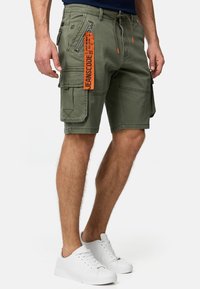 Olive zielone szorty cargo z wieloma kieszeniami, detalem na zamek, regulowanym ściągaczem w talii oraz pomarańczową metką z nazwą marki.