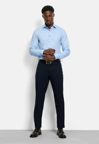 Hellblaues Hemd mit spitzem Kragen, kombiniert mit dunkelblauen Hosen. Ein schwarzer Gürtel und glänzende braune Schuhe runden das Outfit ab.