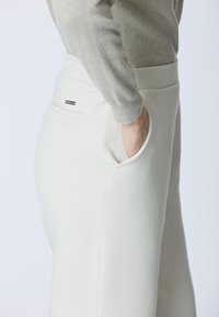 Pantaloni della tuta di colore chiaro con vita elastica, tasche laterali e una piccola etichetta con il marchio sulla tasca posteriore. Tessuto dalla texture liscia.