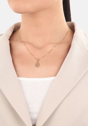 Een gouden ketting met een druppelvormige hanger met een bladerenontwerp, gedragen over een witte top, gecombineerd met een beige blazer.