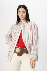 Striped button-up shirt in cream, red, and blue. Features a pocket, worn over a red ribbed top. Brown belt with circular gold accents.