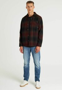 Geruit flanellen shirt in donkerrood en zwart, met een borstzak en knopen, gecombineerd met lichtblauwe jeans en witte sneakers.
