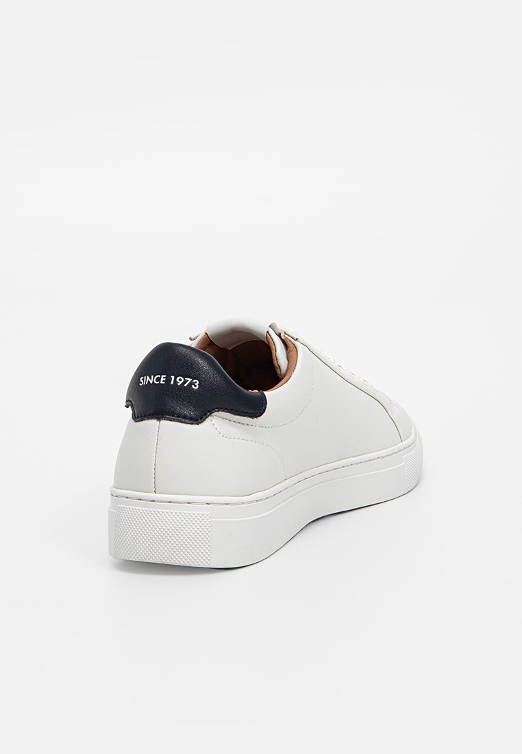 Sneaker en cuir blanc avec un accent de talon en cuir texturé bleu marine. Bout rond, design à lacets, intérieur contrastant en tan, épaisse semelle en caoutchouc blanc.
