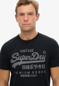 Camiseta de algodón negra con un gran logo gris que lleva el texto "Vintage SuperDry", acompañado de frases menores en asiático e inglés.