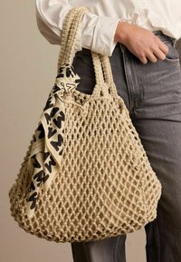 Beige haak tote bag met een gestructureerde open weef en twee gevlochten handvatten. Een gevlochten zwart-wit sjaal is aan een handvat geknoopt.