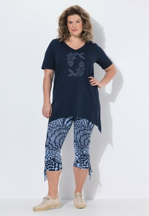 Donna con capelli ricci che indossa una tunica blu navy con design a pesce e leggings corti blu a motivo, in piedi con la mano sui fianchi.
