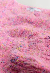 Rozā pūkaina džemperis ar mīkstu tekstūru, kurā ir daudzkrāsaini punkti dzeltenā, zaļā un zilā krāsā. Etiķete redzama netālu no kakla.