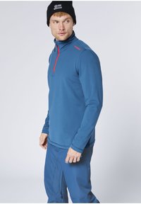 Mann trägt einen blauen Pullover mit Viertel-Reißverschluss und passende Hose, schwarze Mütze mit weißem Logo, blickt über die Schulter vor neutralem Hintergrund.