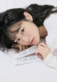 To hvite rør med barrierekrem og lindrende krem, merket med svart tekst, hviler på en hvit overflate. Bakgrunnen har en myk tekstur og en modells hånd.