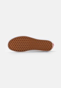 Semelle en caoutchouc brun à motif gaufré d'une basket blanche décontractée avec un petit logo rouge sur la languette du talon.