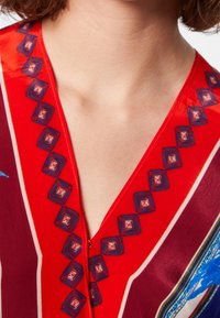 Robe rouge et bordeaux avec un décolleté en V profond, ornée de broderies géométriques bleu et blanc le long du décolleté et d'une texture de tissu lisse.