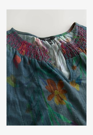 Halenka s květinovým potiskem v tmavě modré barvě, s nařaseným výstřihem v hluboké červené. Měkká, lehká látka s výraznými květinovými vzory a jemnou texturou.