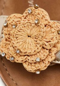 Adorno de flor de paja en color natural con pequeños acentos de cristal en una tira de sandalia metálica color beige, mostrando una textura tejida y un diseño en capas.