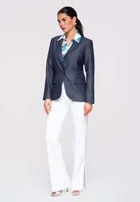 Femme portant un blazer bleu marine sur une chemise fleurie bleue, un pantalon blanc à jambes larges avec des fentes latérales, et des sandales blanches à talons, debout devant un fond clair.