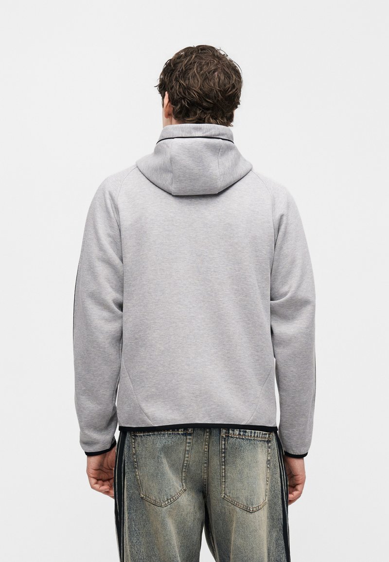 Man draagt lichtgrijze capuchonsweatshirt en vervaagde blauwe jeans, gezien van achteren tegen een effen witte achtergrond.