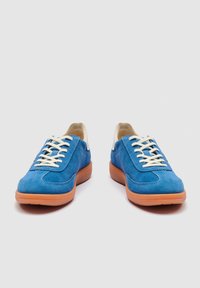 Ζευγάρι μπλε σατέν αθλητικά παπούτσια με λευκά κορδόνια και ανοιχτό καφέ λαστιχένιες σόλες, εμφανίζονται από μπροστά σε απλό φόντο.