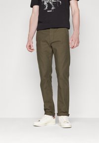 Oliwkowe spodnie z teksturowanej tkaniny o prostym kroju i kieszeniach, zestawione z białymi trampkami i czarnym t-shirtem z nadrukiem.