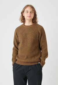 Brauner gestrickter Pullover mit einem strukturierten Muster, Rundhalsausschnitt und gerippten Bündchen, kombiniert mit schwarzen Hosen. Das Design beinhaltet ein Etikett am Saum.