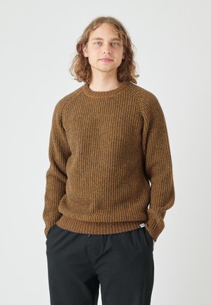 Brauner gestrickter Pullover mit einem strukturierten Muster, Rundhalsausschnitt und gerippten Bündchen, kombiniert mit schwarzen Hosen. Das Design beinhaltet ein Etikett am Saum.