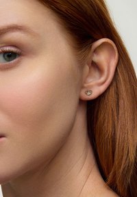 Złote sercowe kolczyki o skomplikowanym wzorze, na tle jasnej karnacji i czerwonych włosów, prezentujące mały, błyszczący detal.