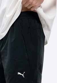 Κοντινό πλάνο ενός ατόμου που σηκώνει λευκό πουκάμισο, φορώντας μαύρα σορτς Puma με ελαστική μέση και εμφανές λογότυπο Puma κοντά στην τσέπη.