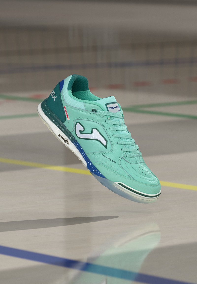 Chaussure de sport d’intérieur turquoise clair avec des accents blancs et bleus, suspendue en l’air au-dessus d’un terrain avec des lignes colorées.