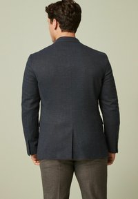 Blazer su misura blu navy con spacco singolo, silhouette aderente e tessuto testurizzato. La parte posteriore presenta spalle strutturate e polsini con bottoni.