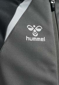 Chaqueta gris con cremallera hecha de un tejido texturizado con un diseño en blanco y negro. Presenta un logo y acentos en los hombros de contraste.