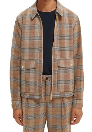 Mężczyzna w jasnobrązowej, czarnej i białej kratowanej kurtce oraz dopasowanych spodniach, z czarną koszulą pod spodem, stoi z rozluźnionymi rękami.