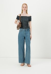Modra široka jeans hlače v kombinaciji z črnim cvetličnim topom brez naramnic. Model drži rjavo torbico in nosi bež sandale s trakovi.