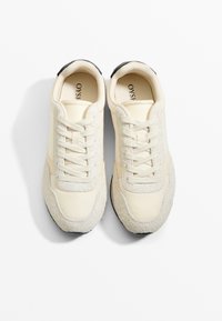 Beiga sneakers med en slät ovandel med mockadetaljer, rund tå och texturerad sula med svart klackhätta. Sex snörhål synliga.