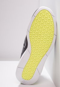 Černá teniska s texturovaným svrškem, bílou podrážkou a výraznou žlutou podrážkou se šestiúhelníkovým vzorem. Vyznačuje se odolnými materiály a moderními designovými prvky.