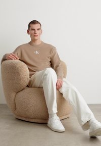 Μπεζ βελούδινη, στρογγυλή καρέκλα με μαλακή υφή, κατειλημμένη από ένα άτομο με κοντομάνικο μπεζ πουκάμισο και λευκά παντελόνια, που κάθεται ήρεμα.