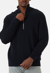 Mann in schwarzem geripptem Reißverschluss-Pullover und hellblauen Jeans, der mit einer Hand den Reißverschluss hochzieht, vor weißem Hintergrund.