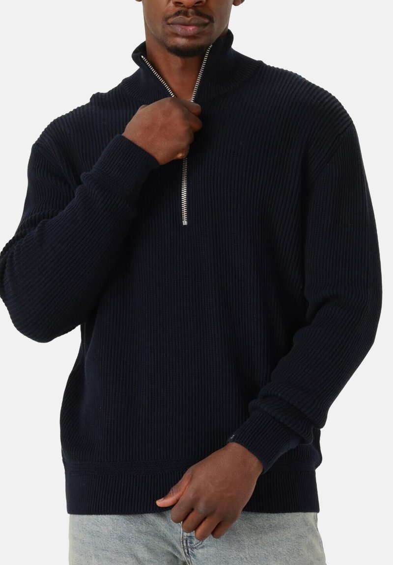 Mann in schwarzem geripptem Reißverschluss-Pullover und hellblauen Jeans, der mit einer Hand den Reißverschluss hochzieht, vor weißem Hintergrund.