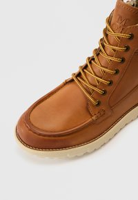 Brun læderankelstøvle med gule snørebånd, rundet tå, polstret krave og lysefarget gummisål. Syninger langs kanten.