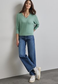 Pull à col en V vert mint avec des poignets côtelés, associé à un jean large bleu et des baskets blanches. Tissu lisse avec un minimum de détails.