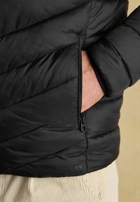 Ruka v zipsovej vrecku čiernej prešívanej bundy, zobrazuje manžetu rukáva a časť béžových kordových nohavíc.