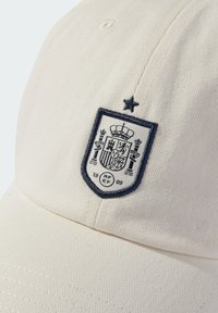 Biała bawełniana czapka z ciemnoniebieskim haftowanym herbem, na którym znajduje się królewska korona oraz różne symbole, w tym gwiazdka nad herbem.