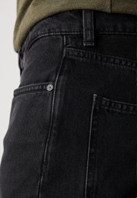 Black denim jeans featuring a textured surface, visible stitching, a small button on the front pocket, and a close-fitting design.