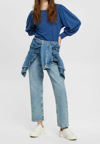 Žena oblečená v modrém topu s nafouknutými rukávy a světlých modrých džínách, s džínsovou bundou uvázanou kolem pasu, stojící s rukou na boku.