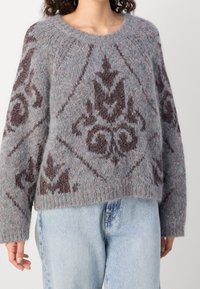 Žena v načechraném šedém svetru s tmavě hnědými zdobenými vzory a světlými modrými džíny, stojící na jednobarevném pozadí.