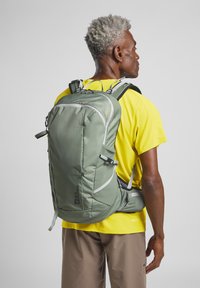 Rugzak in gedempt groene stof, met meerdere compartimenten, verstelbare schouderbanden en reflecterende accenten voor zichtbaarheid.