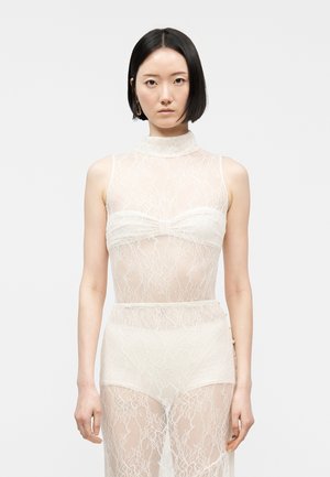 Egyenes fekete hajú nő ujjatlan, áttetsző fehér csipkeruhát visel, mely rétegezve van hozzáillő, magas derekú bugyival és pánt nélküli melltartóval.