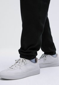 Weiße Leinenschuhe mit dicken Sohlen, Schnürsenkeln und subtiler Markenkennzeichnung, kombiniert mit schwarzen Jogginghosen, die an den Knöcheln gerafft sind, und hellblauen Socken.