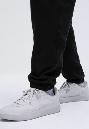 Witte canvas sneakers met dikke zolen, veters en subtiele branding, gedragen met zwarte joggers die bij de enkels zijn verzameld en lichtblauwe sokken.