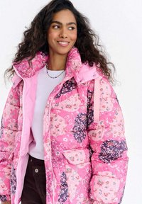 Różowa pikowana kurtka puchowa z kwiatowymi i paisley wzorami, wysokim kołnierzem i kieszeniami. Projekt łączy odcienie fioletu i różu.