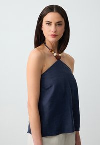 Top halter blu in lino, caratterizzato da una vestibilità semplice e rilassata, con un laccio al collo e dettagli decorativi in legno attorno al colletto.